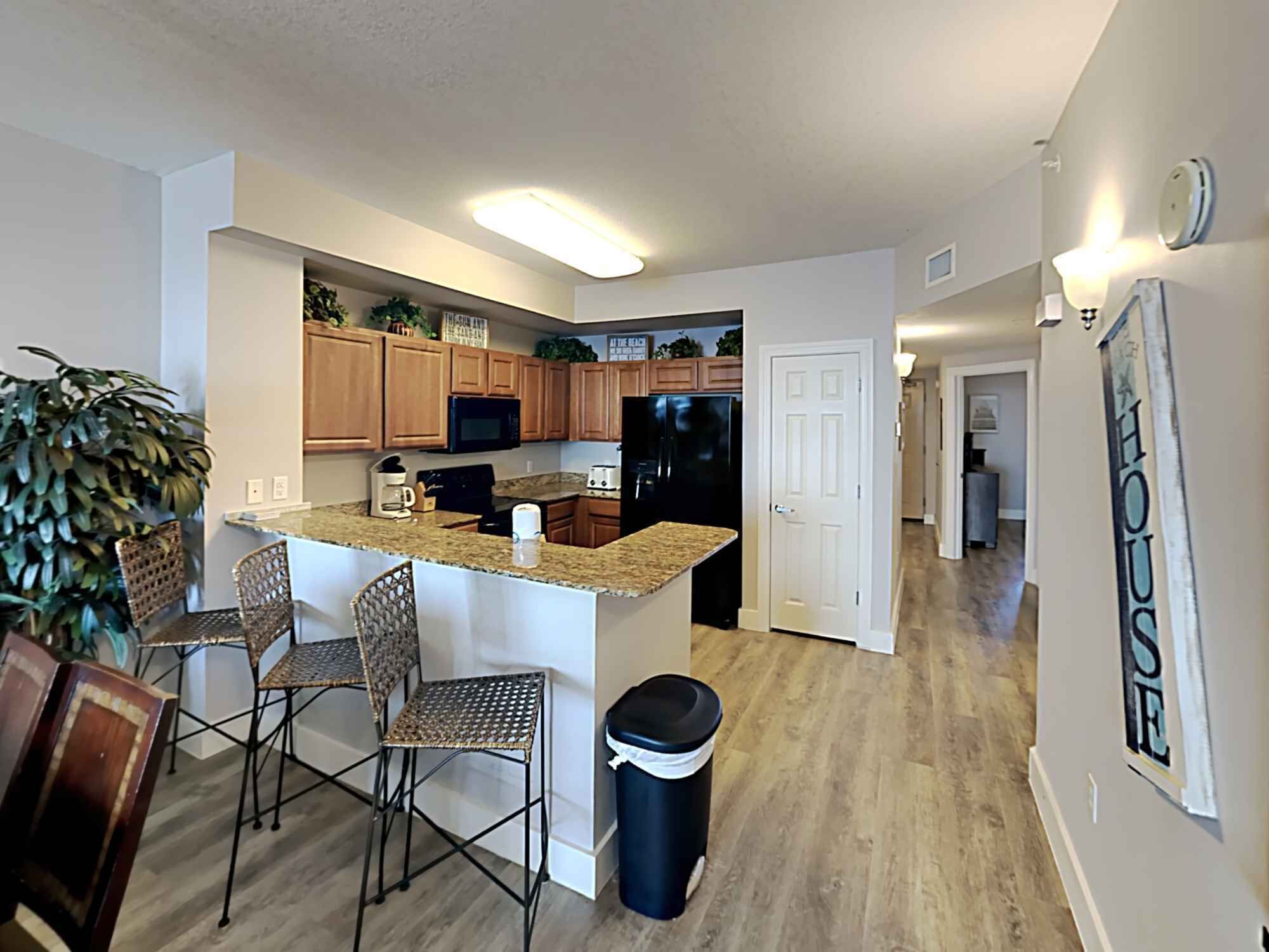 A modern kitchen with wooden cabinets, a black refrigerator, a microwave, and a granite countertop. There are wicker chairs and a trash can.