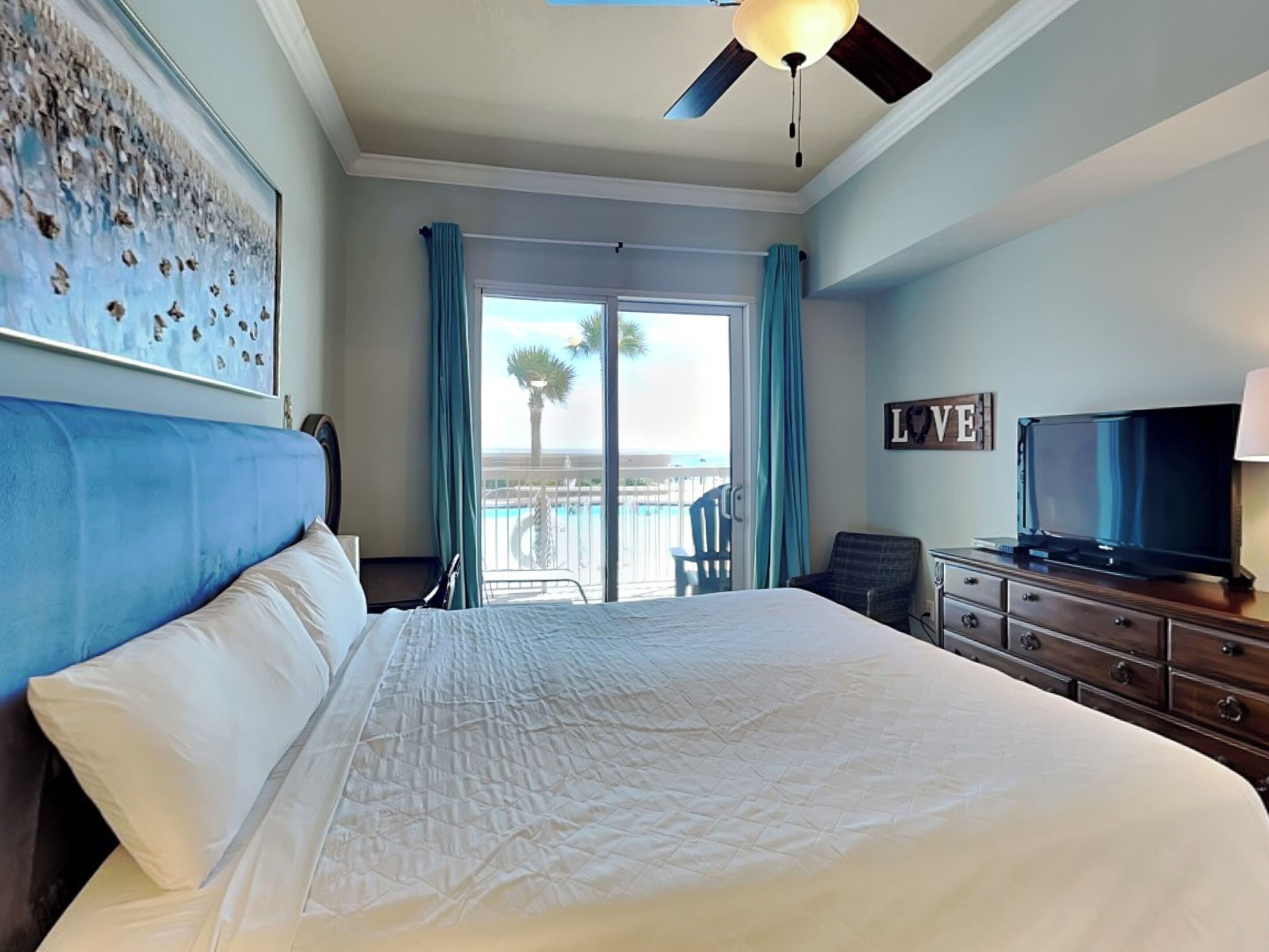 A hotel room with a large bed, a television, a desk, and a view of the pool and palm trees.