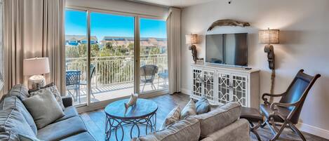 Un salón amplio con un sofá grande, una mesita redonda y un televisor. Hay un balcón con vistas al paisaje.
