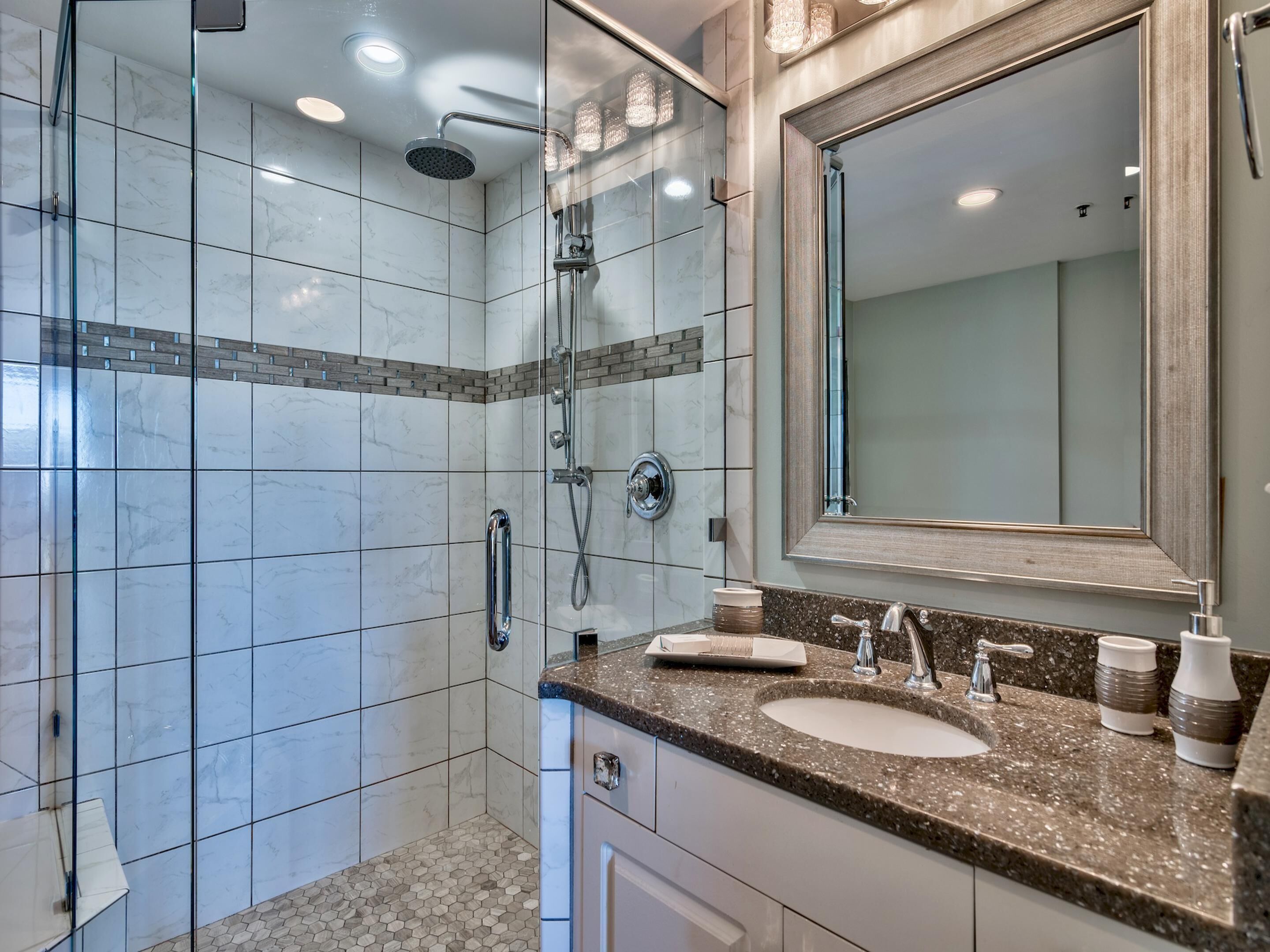 Un baño moderno con ducha envuelta en vidrio, un espejo grande y una encimera de granito con un lavabo y dos tazas.