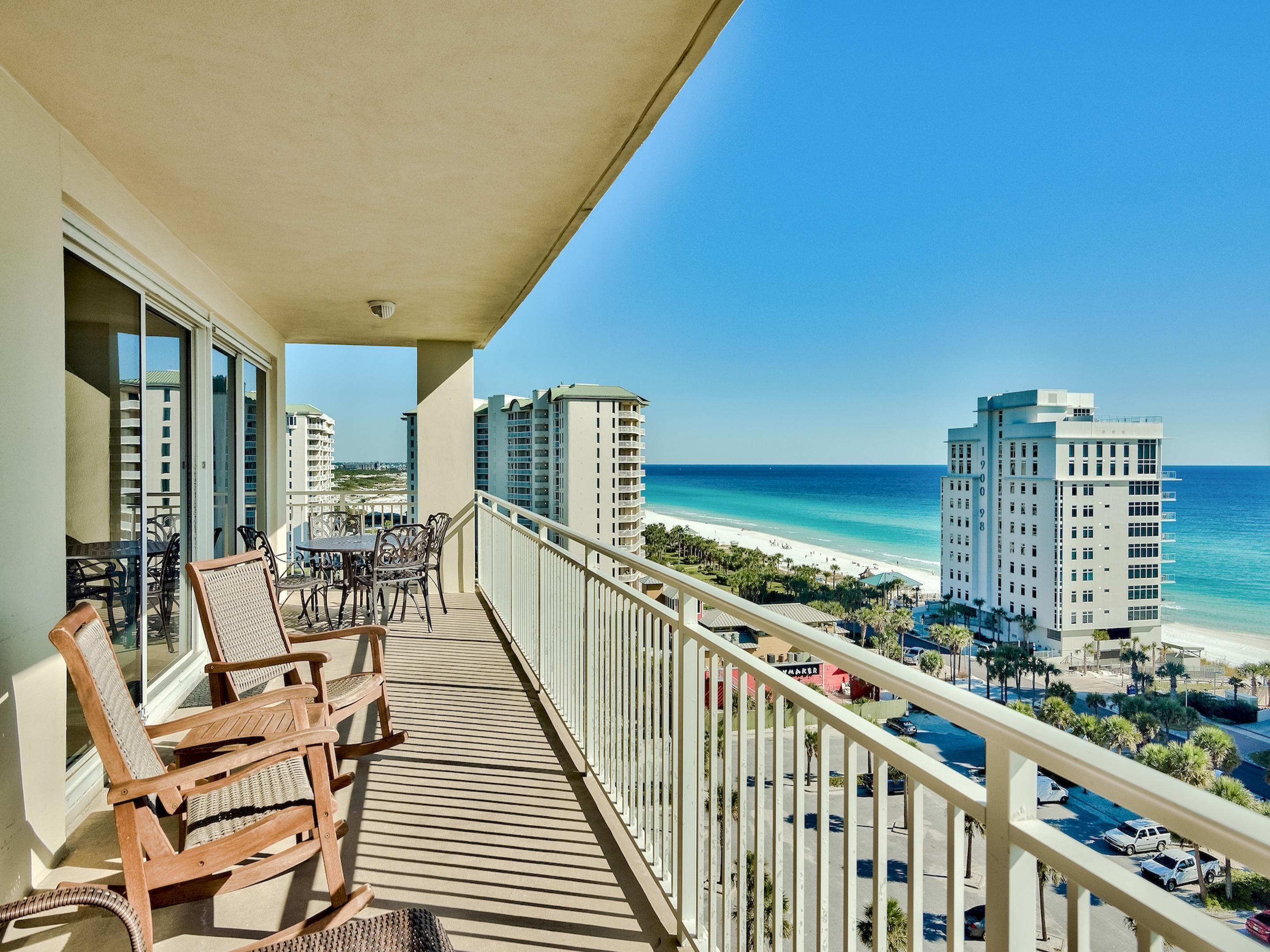 Un balcone con sedie e tavolo in legno, con vista su una spiaggia, edifici alti e cielo azzurro intenso.