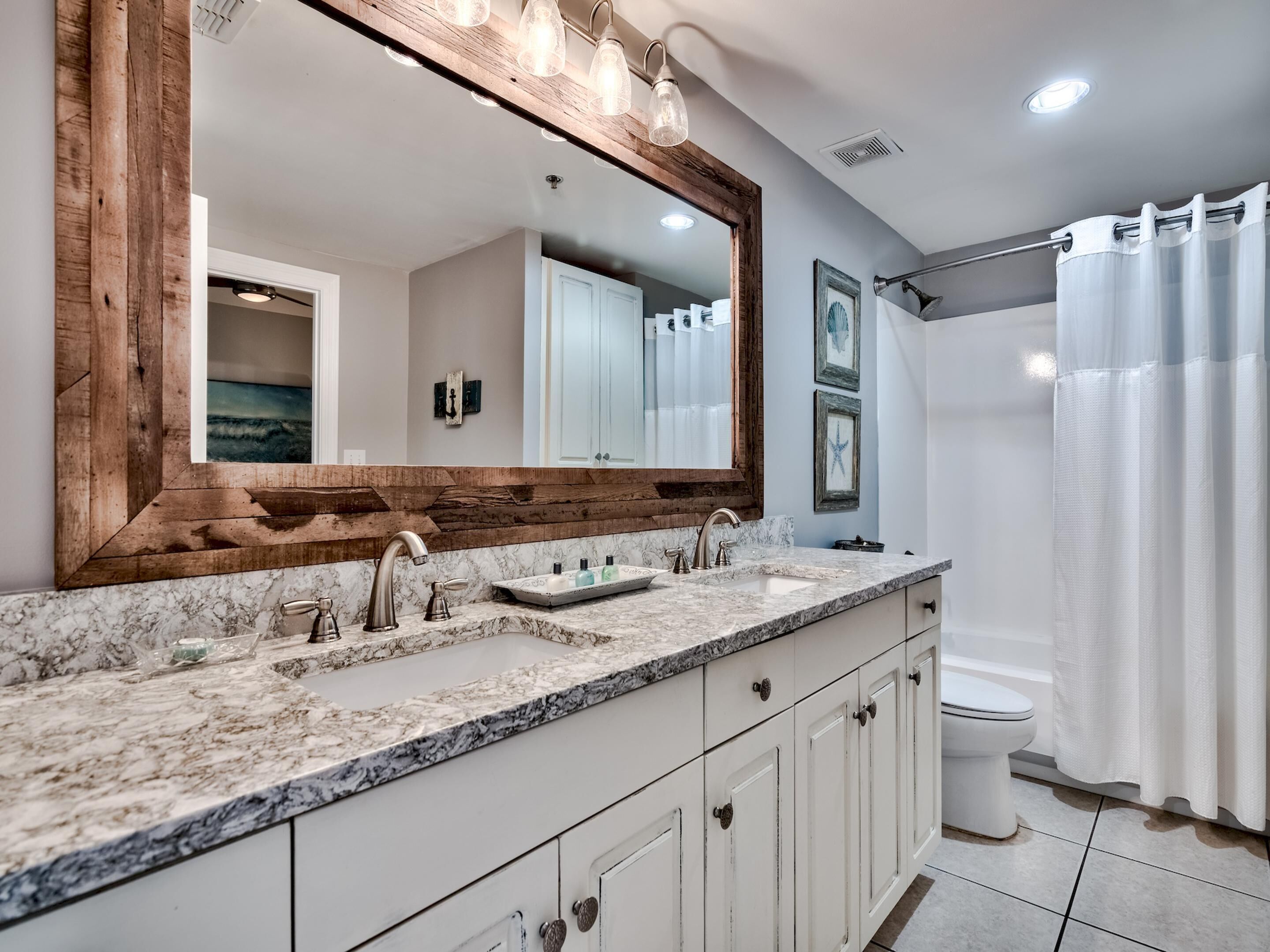 A bathroom with a large mirror, marble countertop, white cabinets, and a shower curtain.