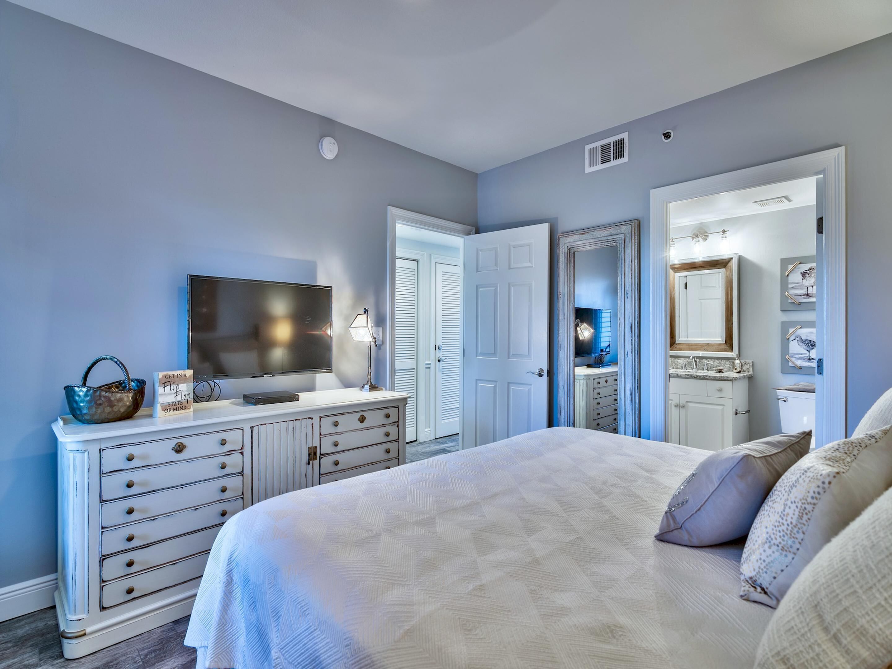 A bedroom with a bed, dresser, vanity, and a television.