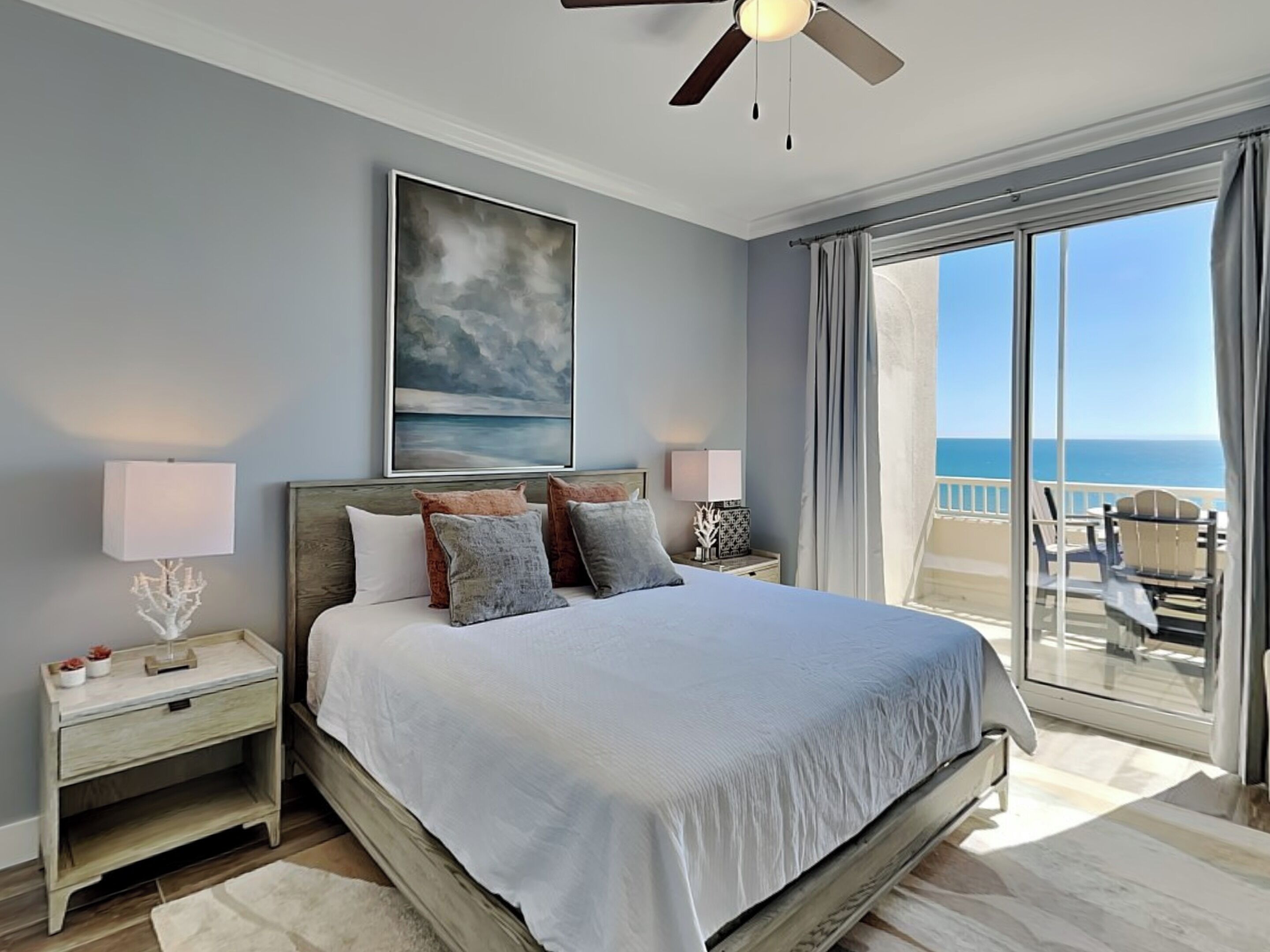 A bedroom with a large bed, a ceiling fan, a painting, and a view of the ocean through a sliding glass door.