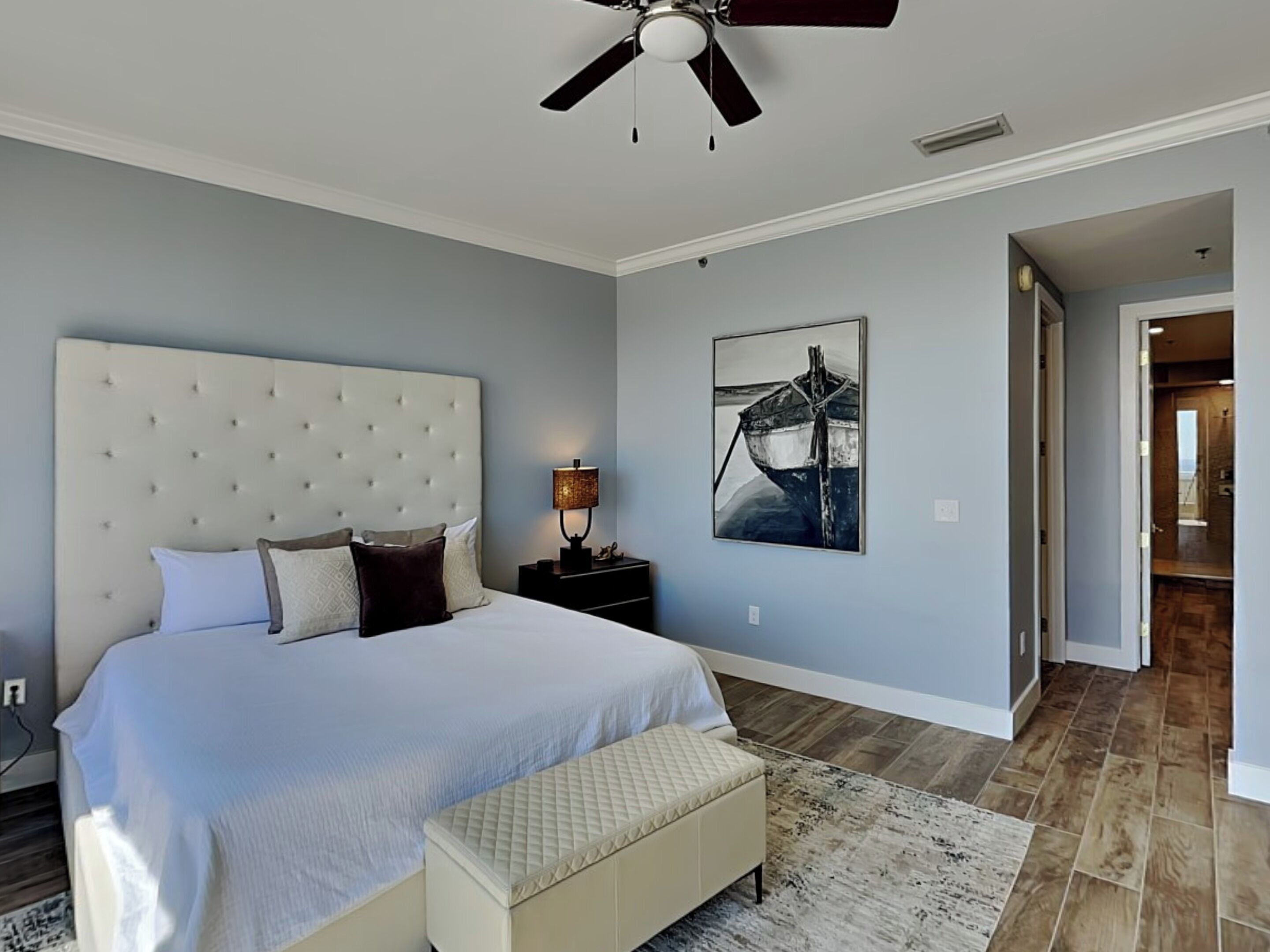 A bedroom with a large bed, a ceiling fan, a nightstand, a bedside table with a lamp, and a framed picture on the wall.