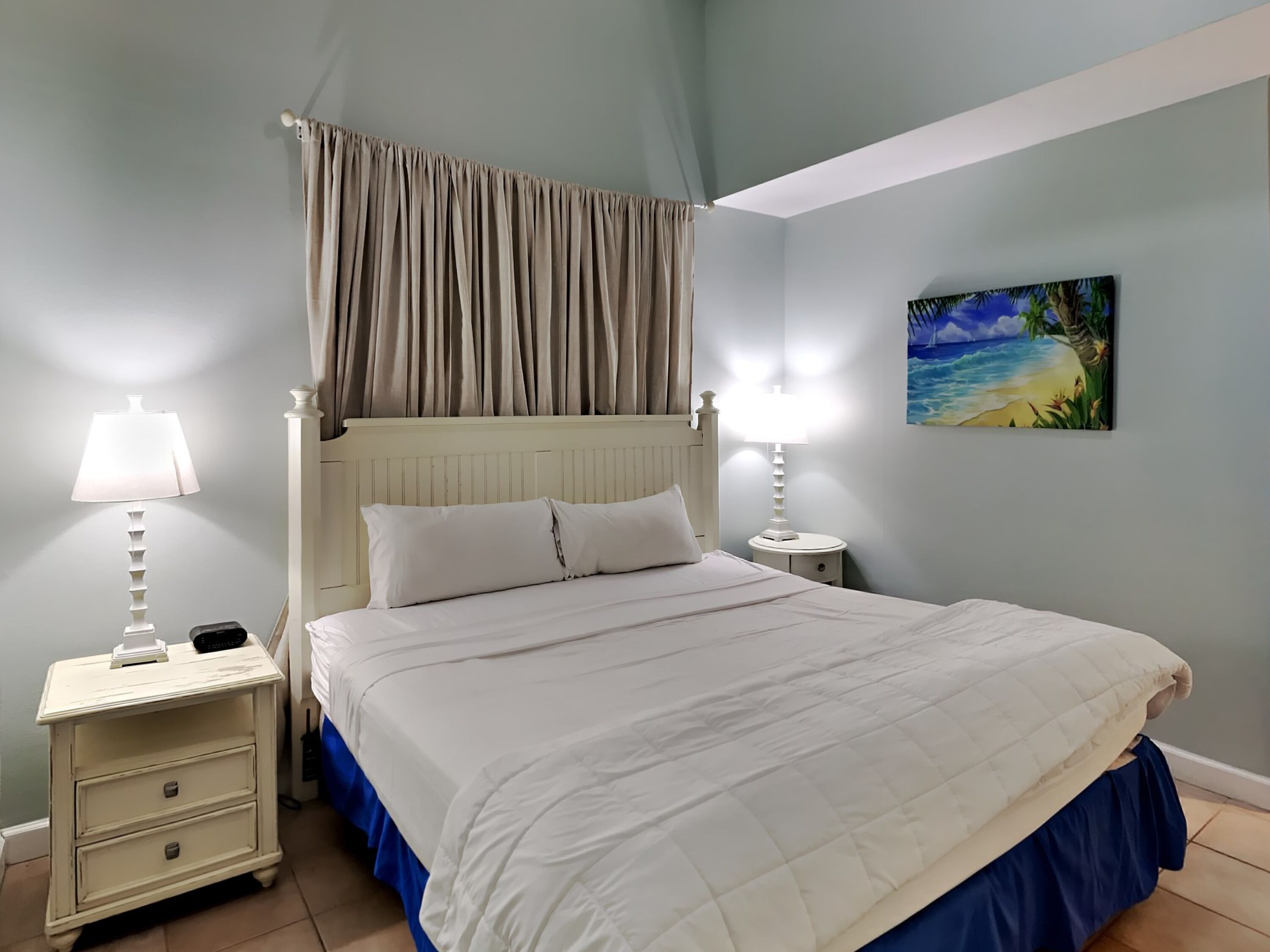 A bedroom with a bed, two bedside tables with lamps, a painting on the wall, and a window with curtains.
