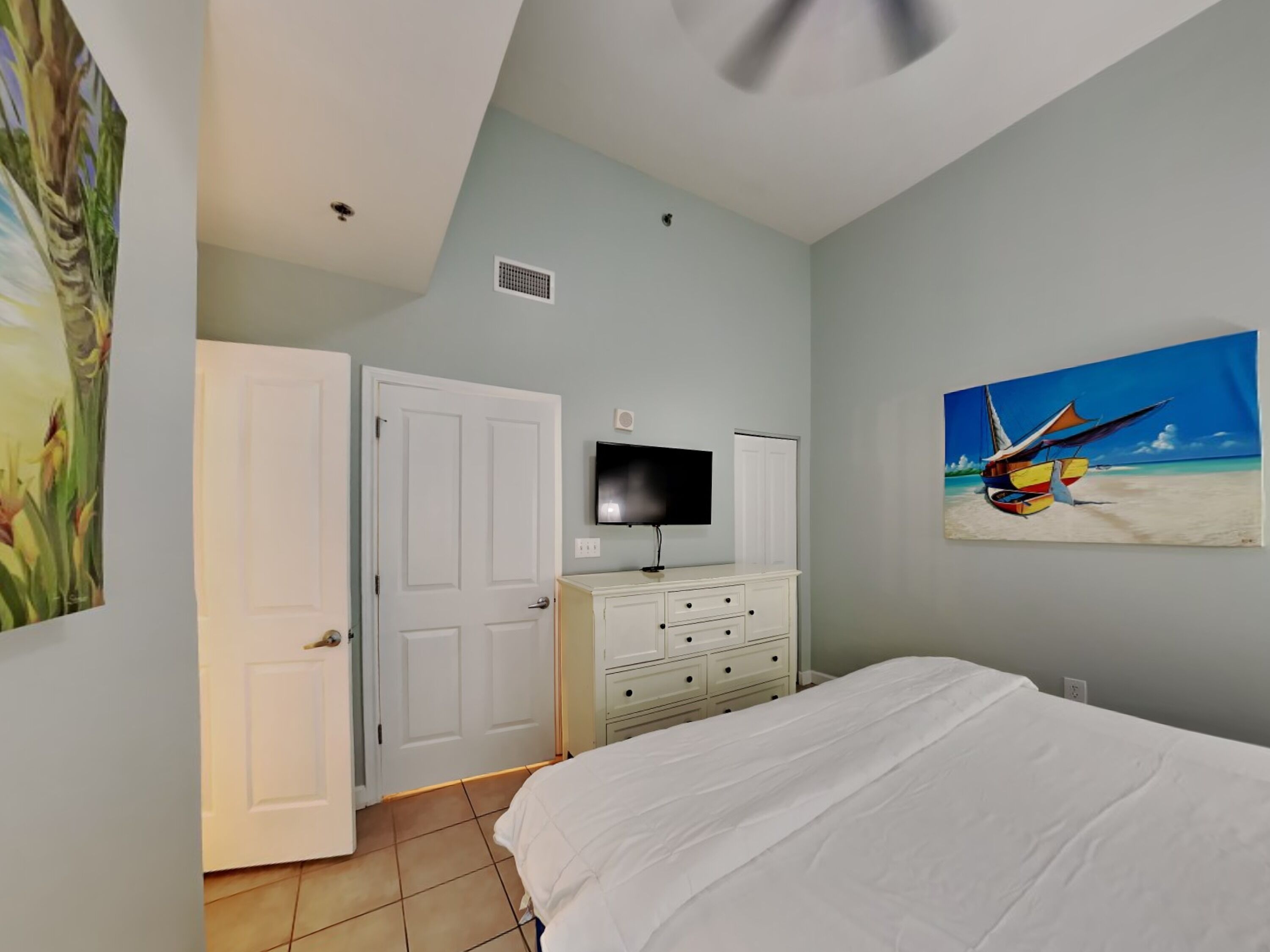A bedroom with a bed, a TV, a dresser, and a painting of a boat on a beach.