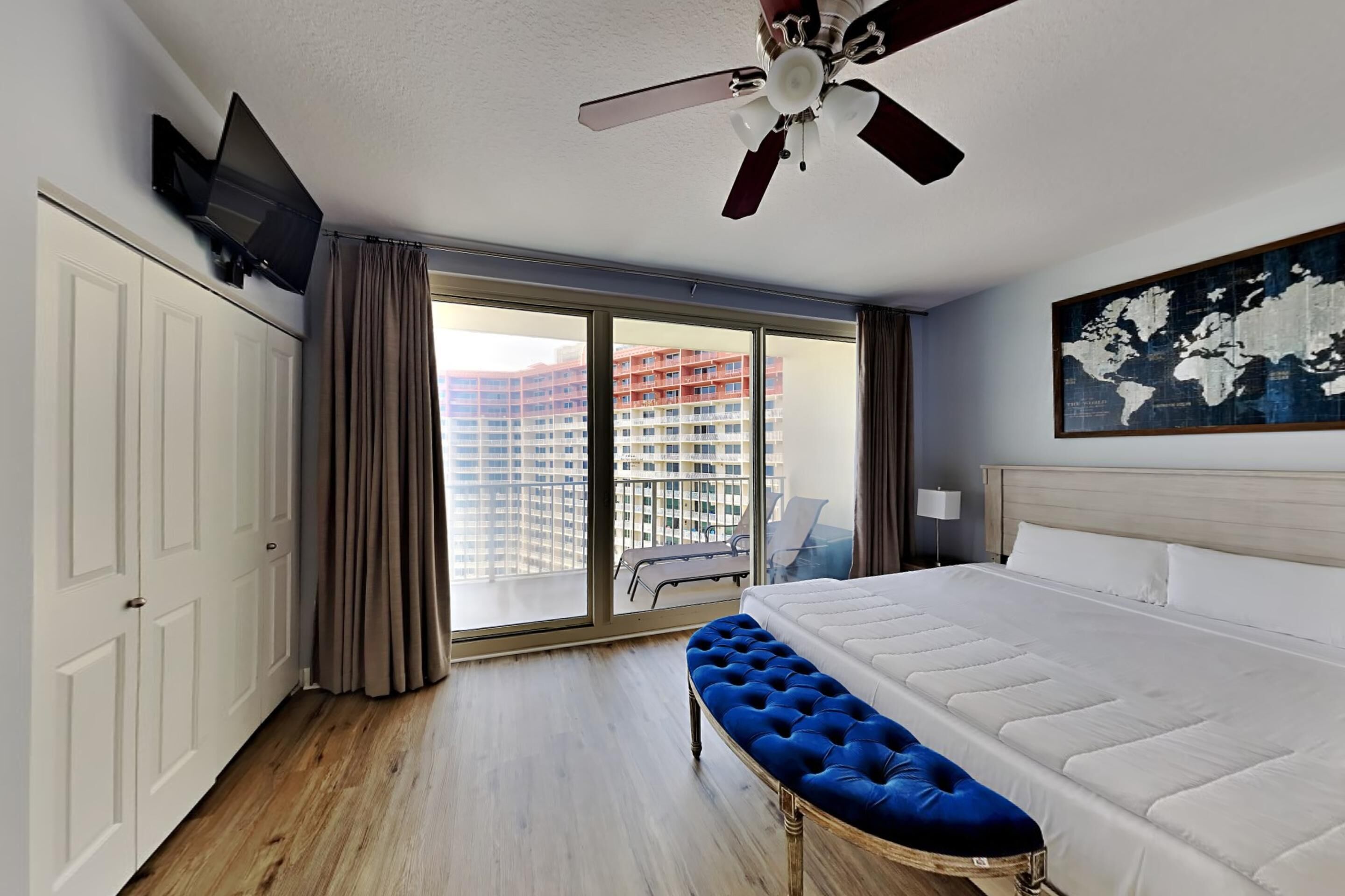 Ein Hotelzimmer mit Bett, einer blauen Bank, einem Deckenventilator, einem Fernseher und einem großen Fenster mit Blick auf hohe Gebäude.