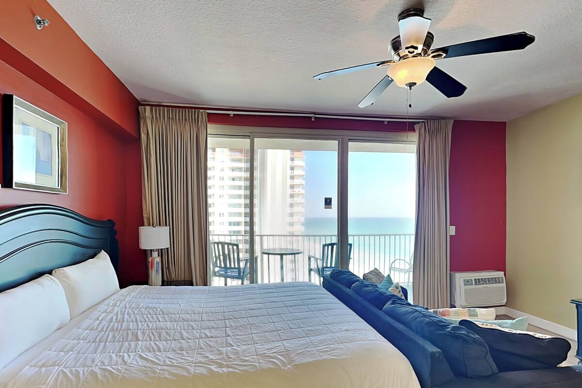A hotel room with a large bed, a ceiling fan, a balcony with a view of the ocean, and a window with curtains.