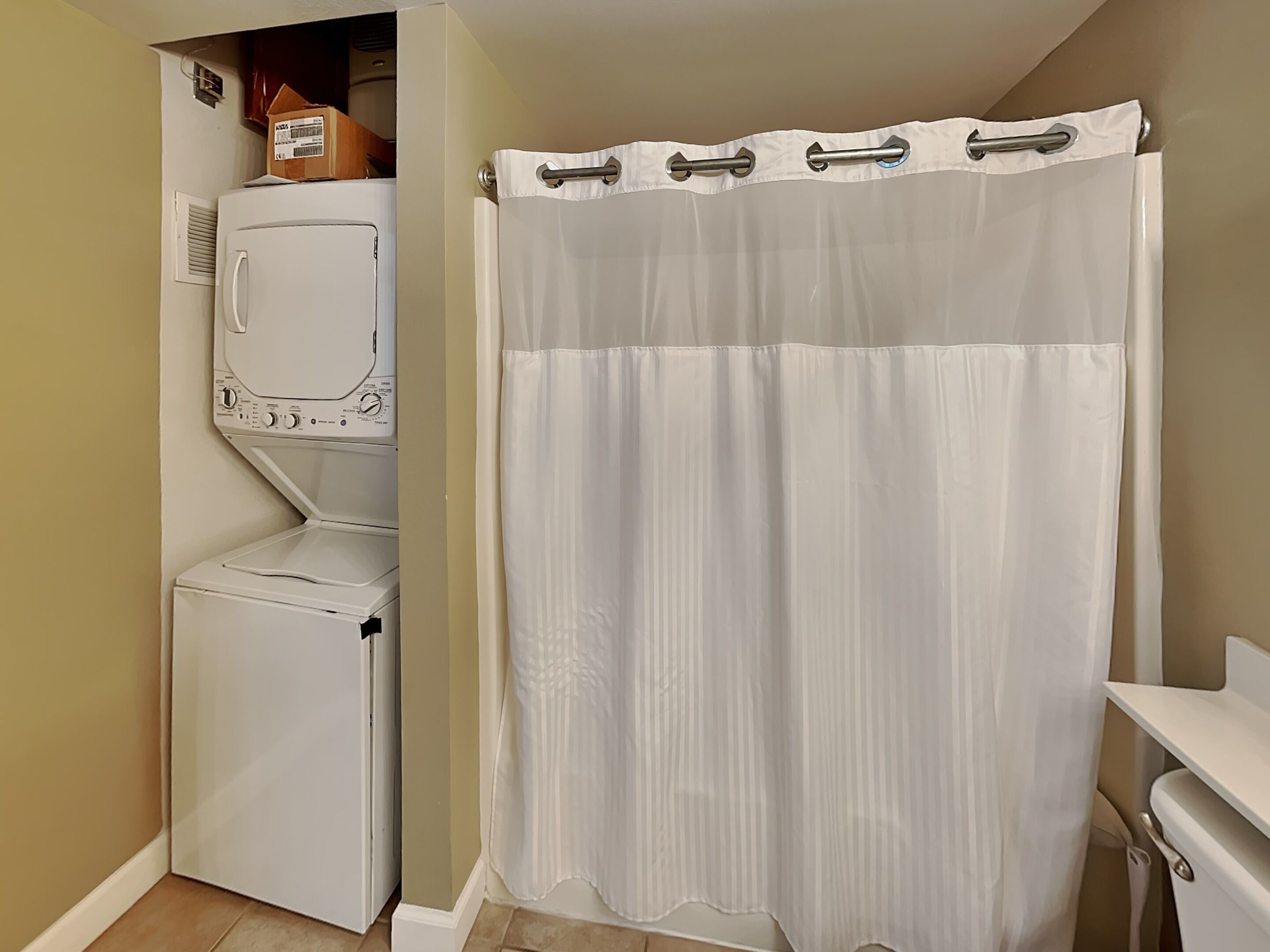 Une buanderie équipée d’un lave-linge et d’un sèche-linge, d’un rideau de douche et d’un petit meuble sur lequel est posée une boîte.