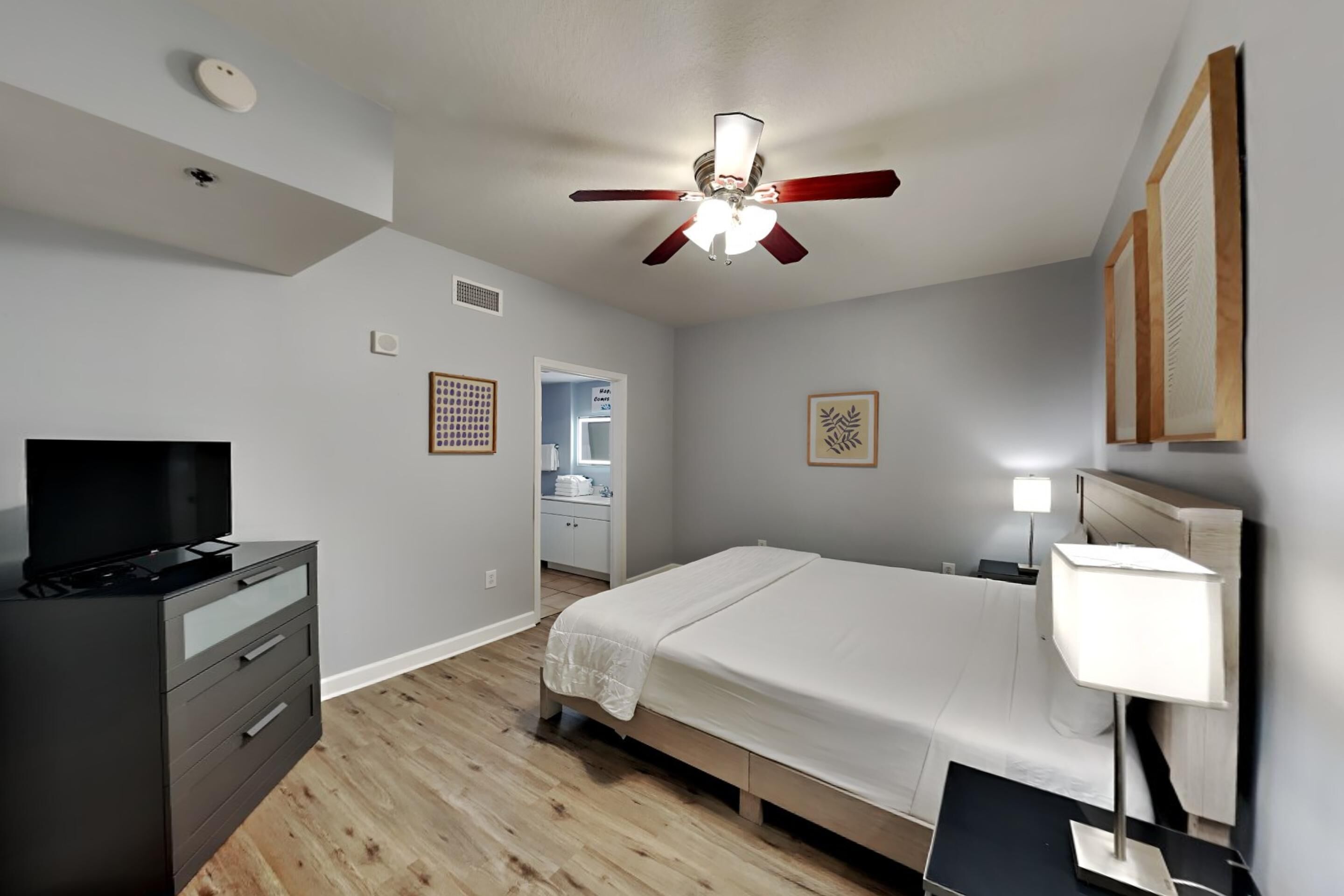 A bedroom with a bed, a TV, a desk, a ceiling fan, and a nightstand with a lamp.