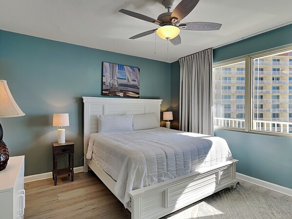 Une chambre à coucher avec un lit, un ventilateur de plafond, une table de chevet avec une lampe, un tableau au mur et une fenêtre donnant sur des immeubles.