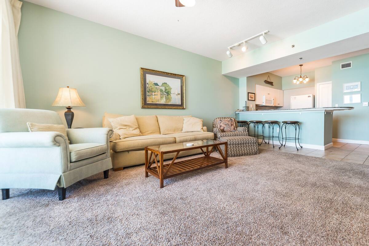 A living room with a sofa, armchairs, a coffee table, and a kitchen area with a bar and stools.
