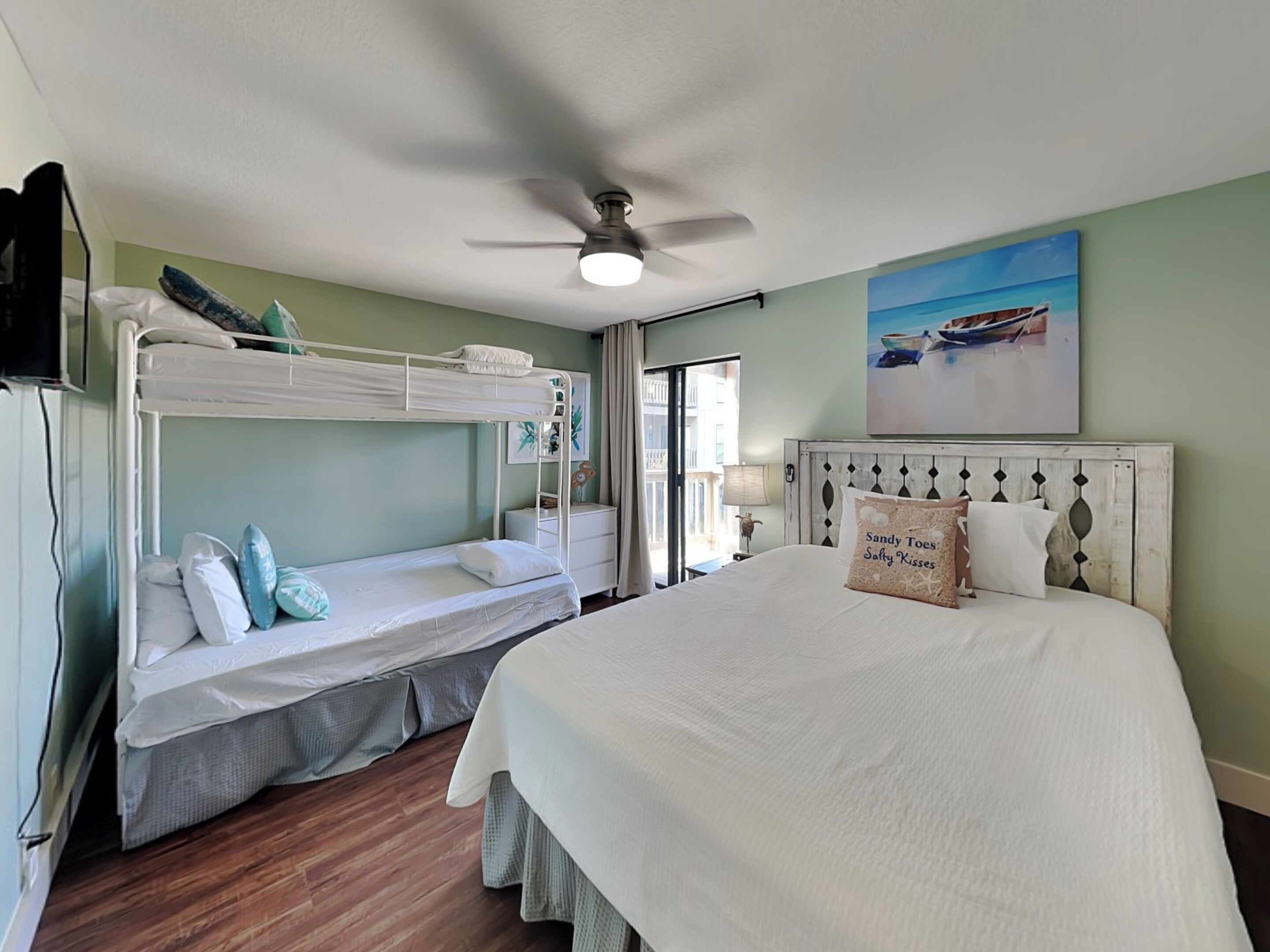 Habitación de hotel con literas, cama grande, ventilador de techo, pintura de barcos y vista al mar.