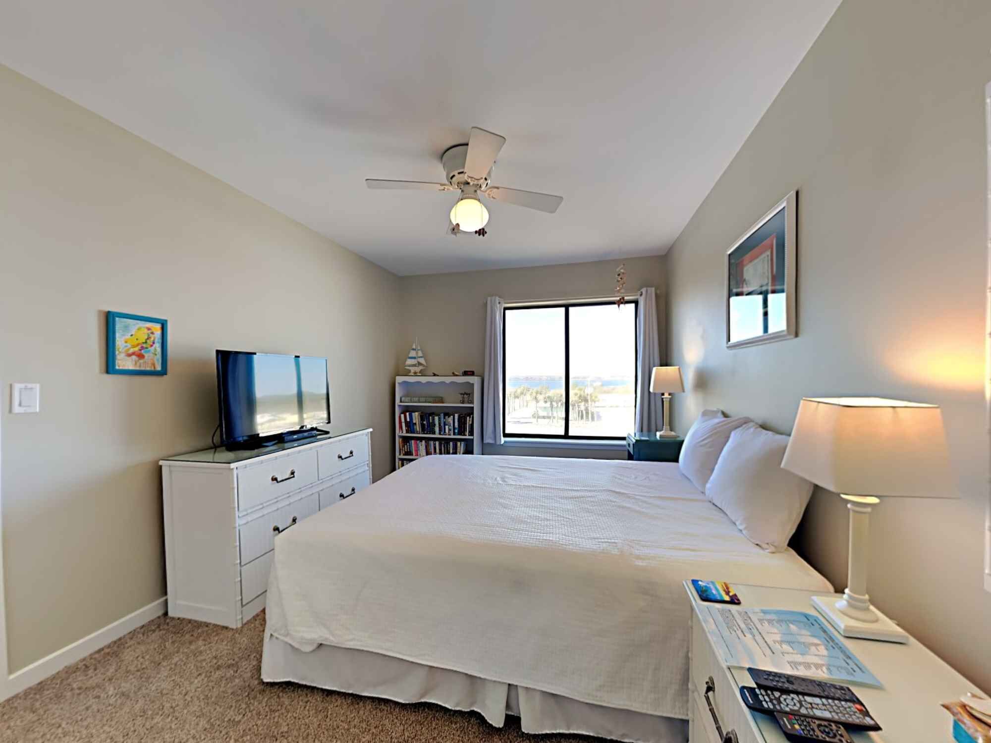 A hotel room with a bed, a television, a bookshelf, a window with a view, and a ceiling fan.