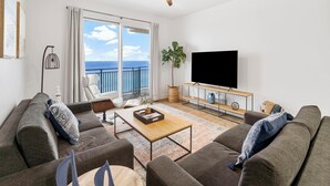 Ein Wohnzimmer mit großem Fernseher, zwei Sofas, einem Couchtisch und einem Balkon mit Meerblick.