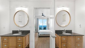 Ein modernes Badezimmer mit zwei Waschbecken, Holzausbau und großen Spiegeln. Durch eine geöffnete Tür sind ein Bett und ein Balkon mit Blick zu sehen.