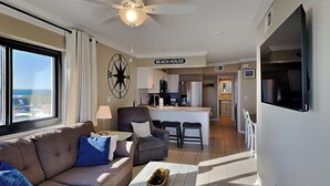 Un salon moderne avec un canapé, une table basse et un ventilateur de plafond. On aperçoit à l’arrière-plan un coin cuisine avec une enseigne indiquant « MAISON DE PLAGE ».
