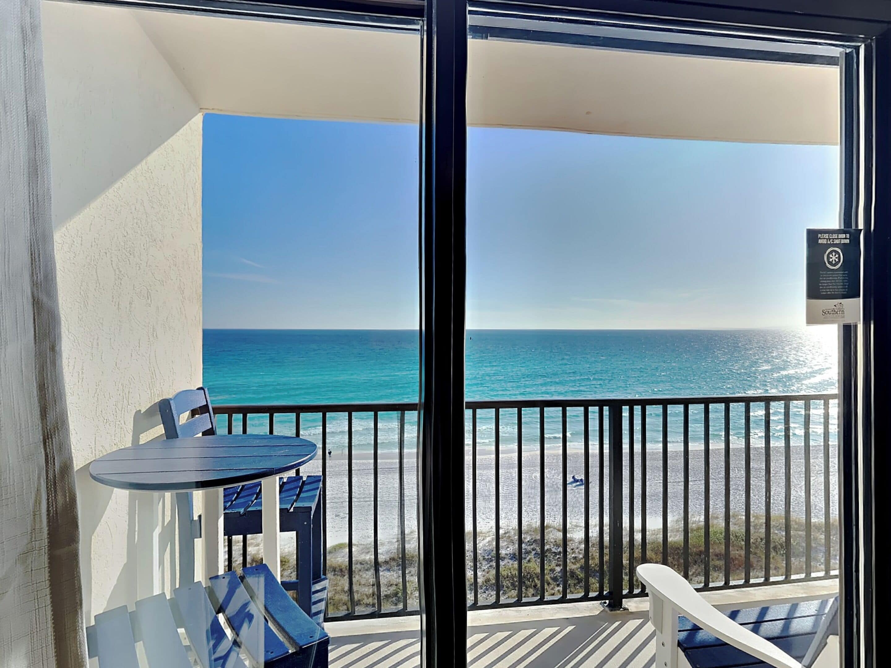 A balcony with a table and chairs, overlooking a beach with clear blue water and a sandy shore.