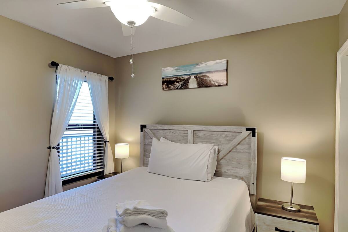 A neatly made bed with white linens, a wooden headboard, and a painting of a beach scene above it.