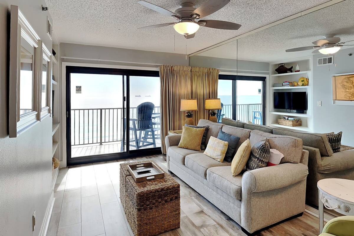 A spacious living room with a large sofa, a coffee table, and a ceiling fan. There is a balcony with a chair and a view of the ocean.