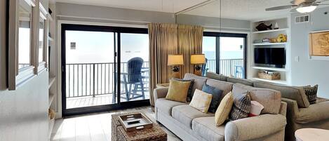 A spacious living room with a large sofa, a coffee table, and a ceiling fan. There is a balcony with a chair and a view of the ocean.