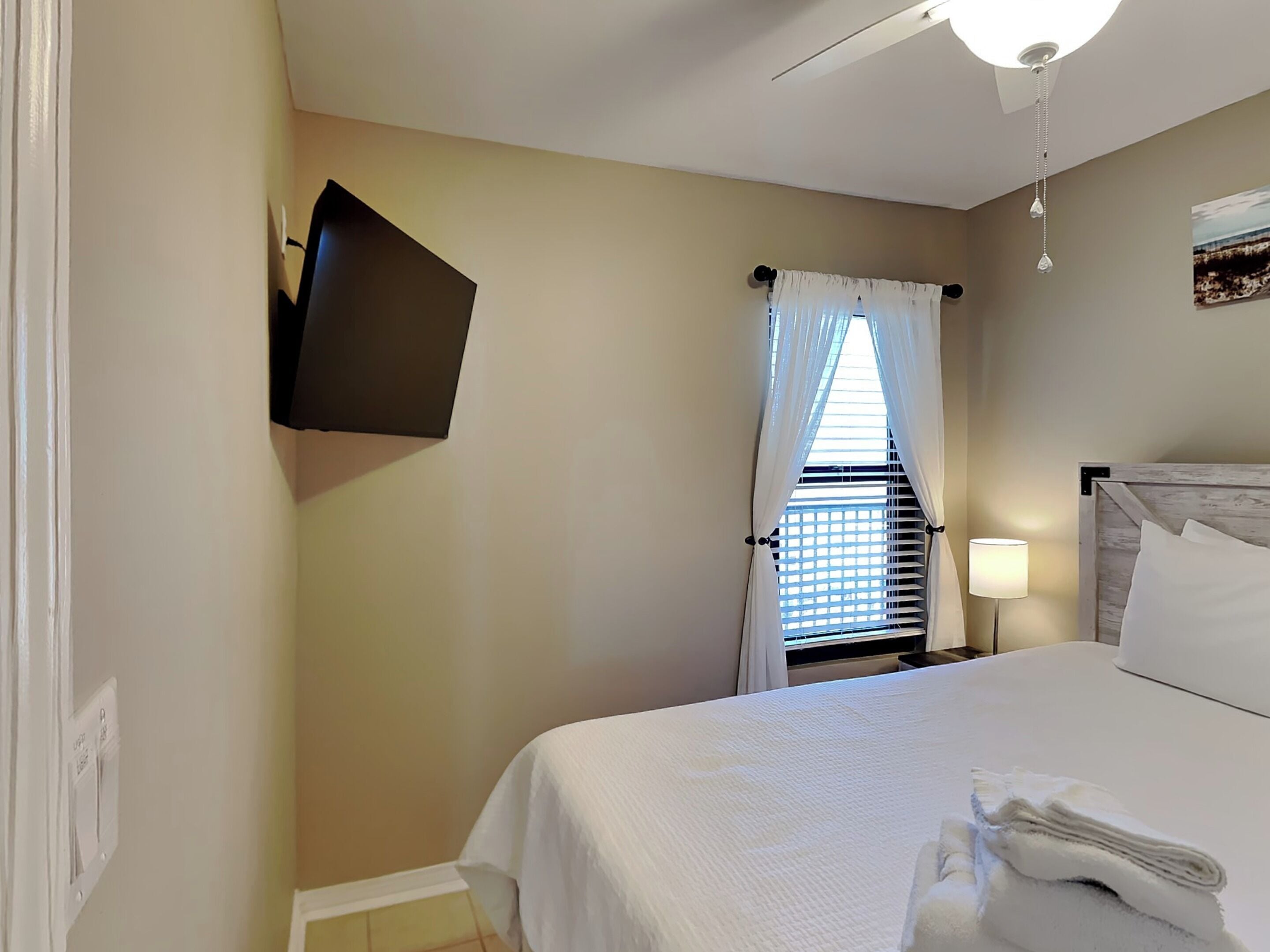 Una habitación de hotel con cama, televisor de pantalla plana montado en la pared, ventana con cortinas blancas, ventilador de techo y lámpara de noche.