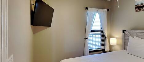 Una habitación de hotel con cama, televisor de pantalla plana montado en la pared, ventana con cortinas blancas, ventilador de techo y lámpara de noche.
