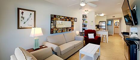 A living room with a sofa, a red armchair, a coffee table, a ceiling fan, and a TV mounted on the wall.