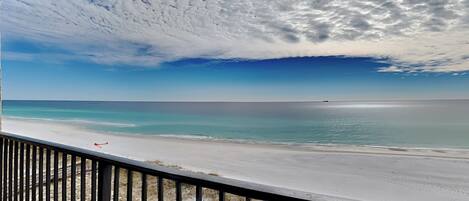 Vista a la playa desde un balcón con barandilla, cielo despejado y mar tranquilo.