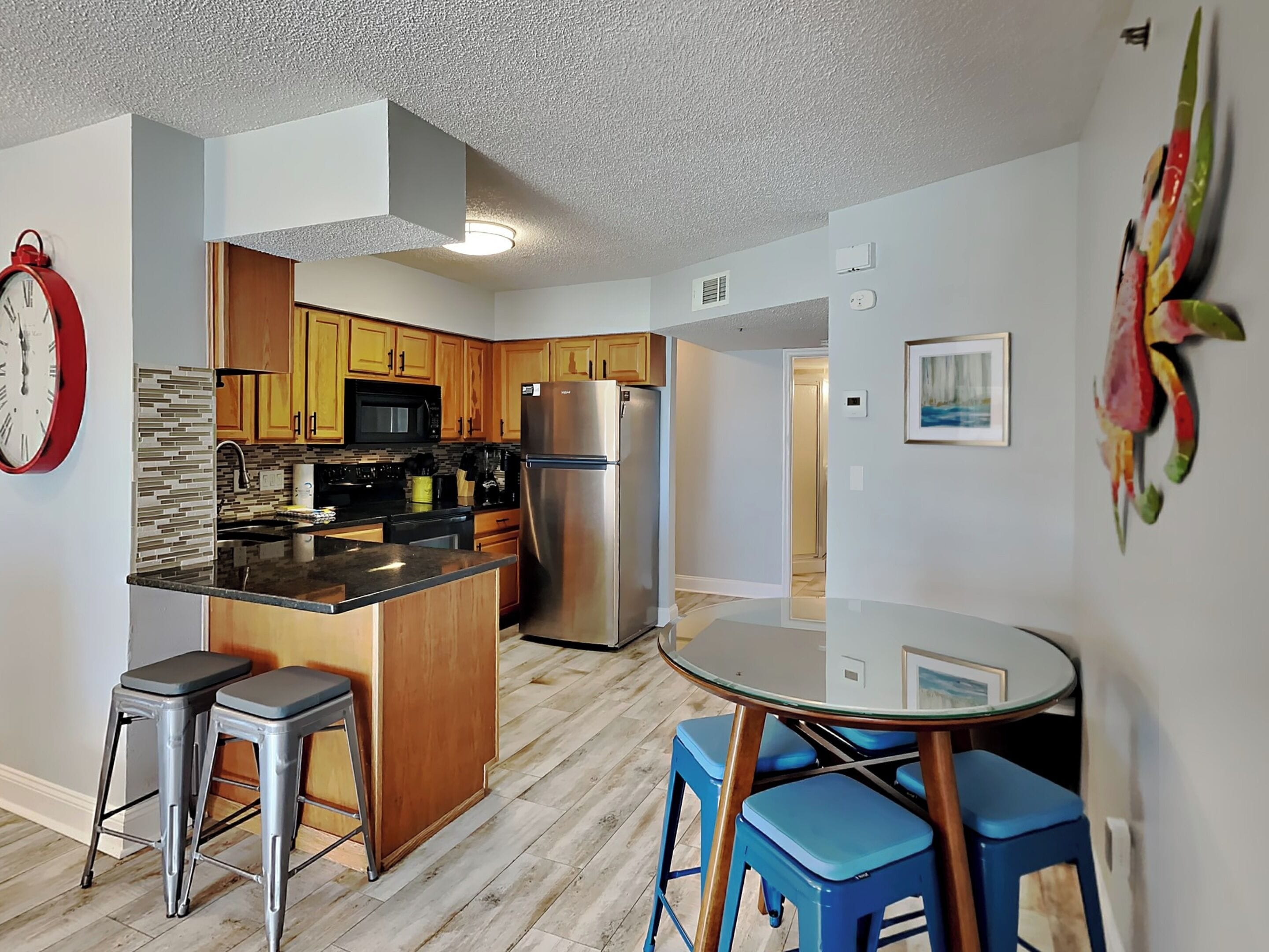 A modern kitchen with wooden cabinets, a stainless steel refrigerator, and a built-in microwave. There are two stools and a glass table with blue chairs.