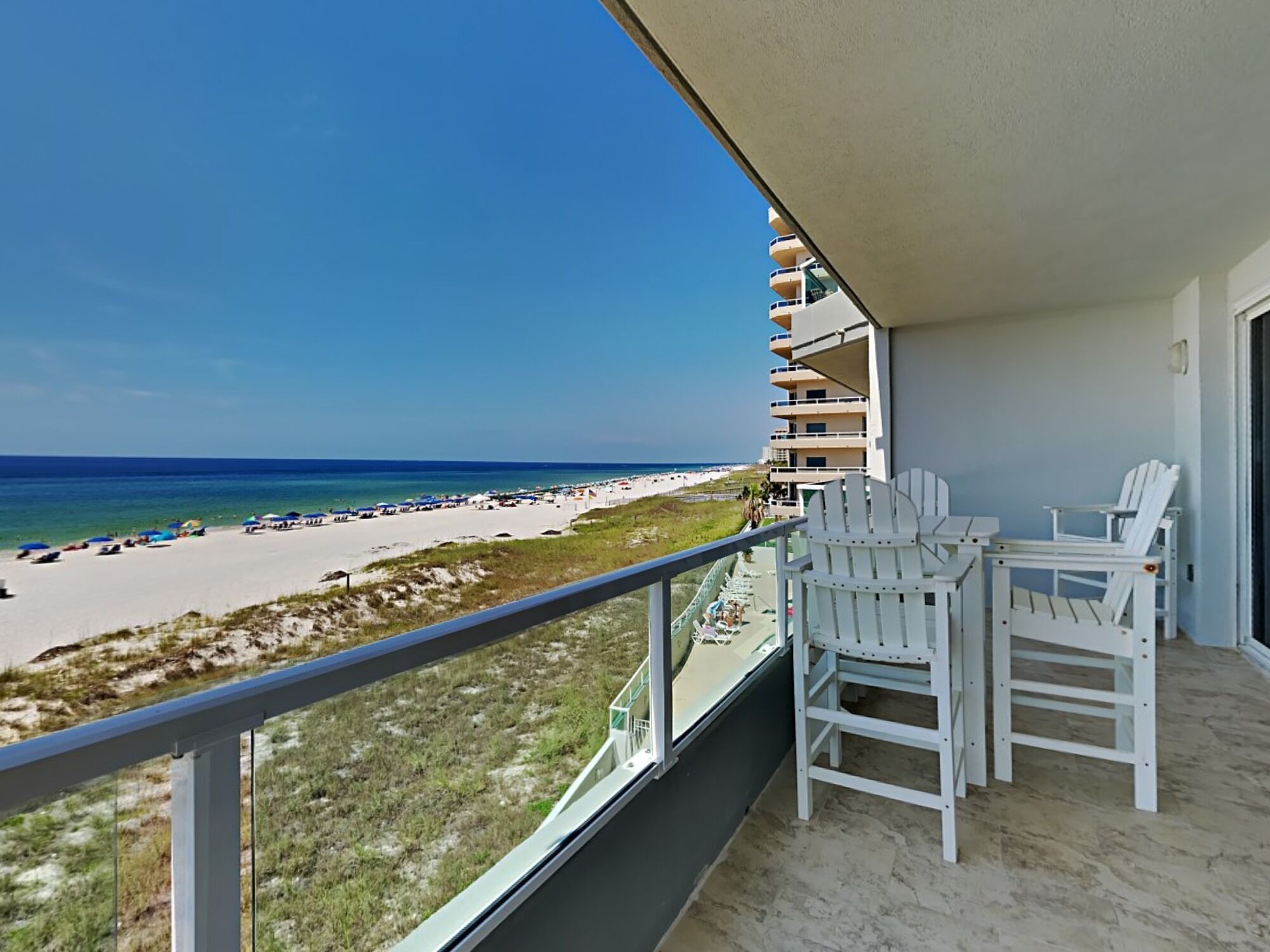Een balkon met witte Adirondack-stoelen, uitzicht op het strand en een helderblauwe lucht.