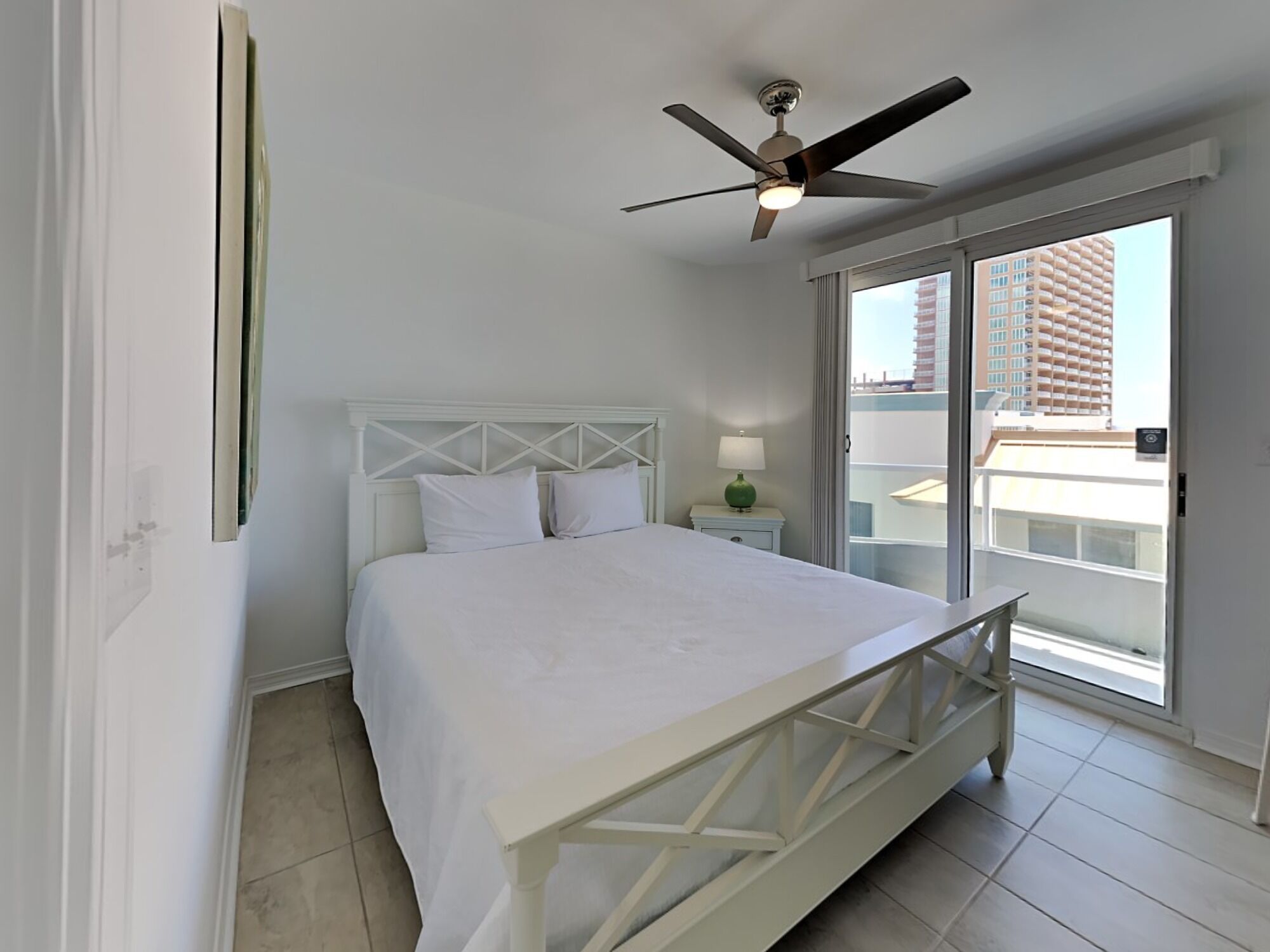 A hotel room with a bed, a ceiling fan, a nightstand with a lamp, and a view of the city through a large window.