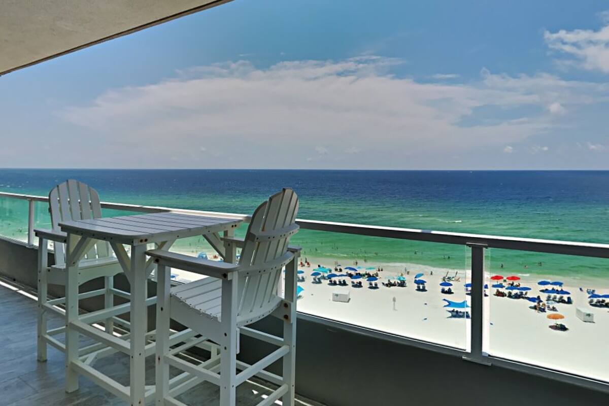 A balcony with two Adirondack chairs facing a beach with umbrellas and people.