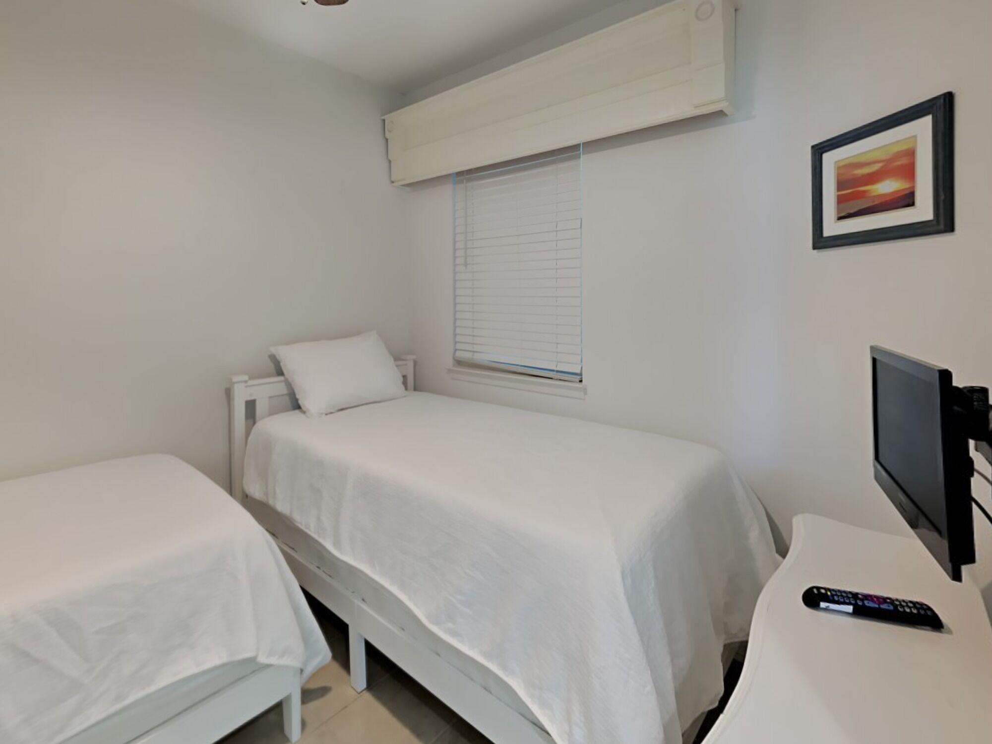 A small, clean room with two single beds, a TV, and a window with blinds.
