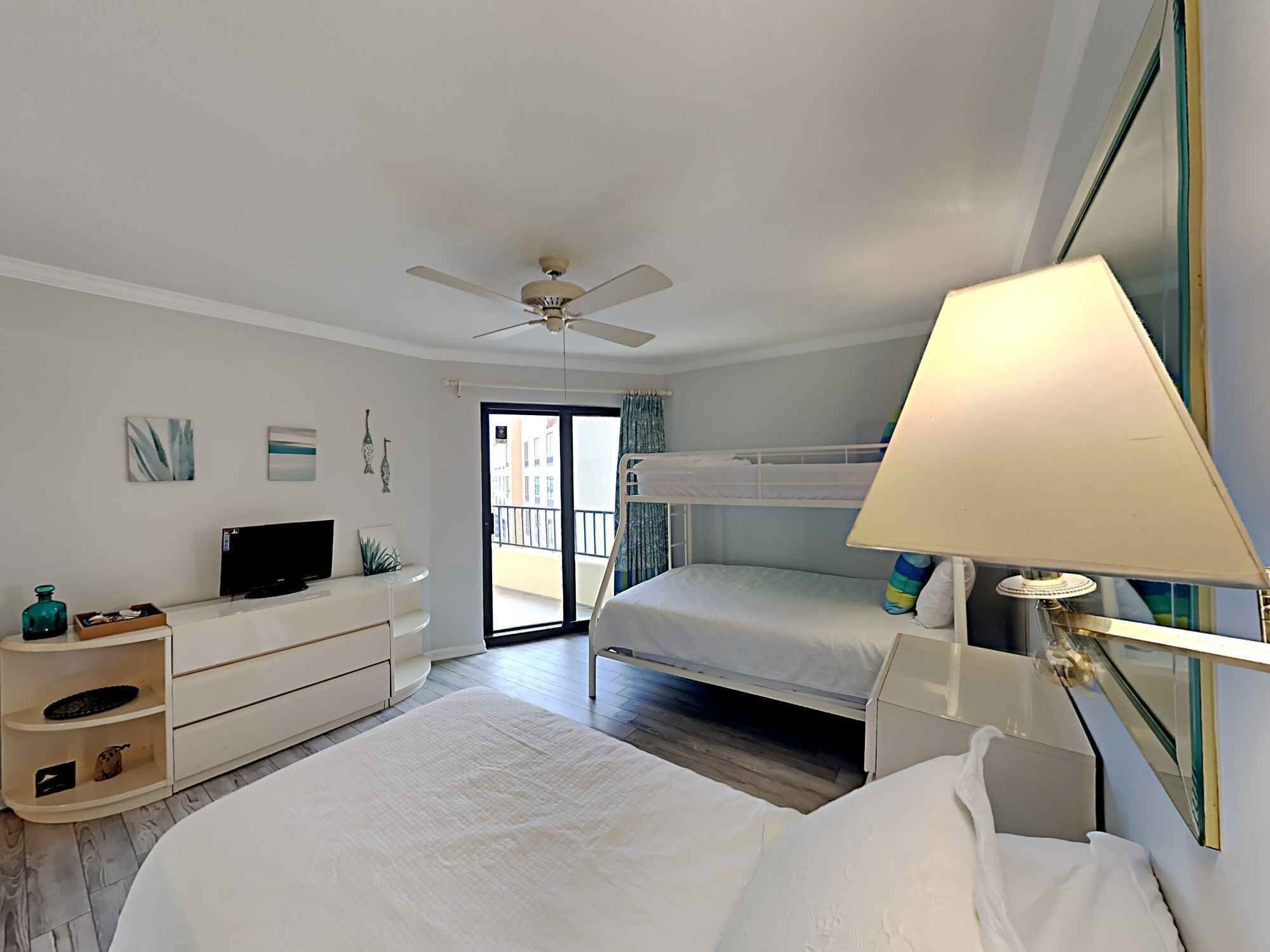 Una camera d'albergo con un letto, una scrivania, una TV, una ventola a soffitto e un balcone.