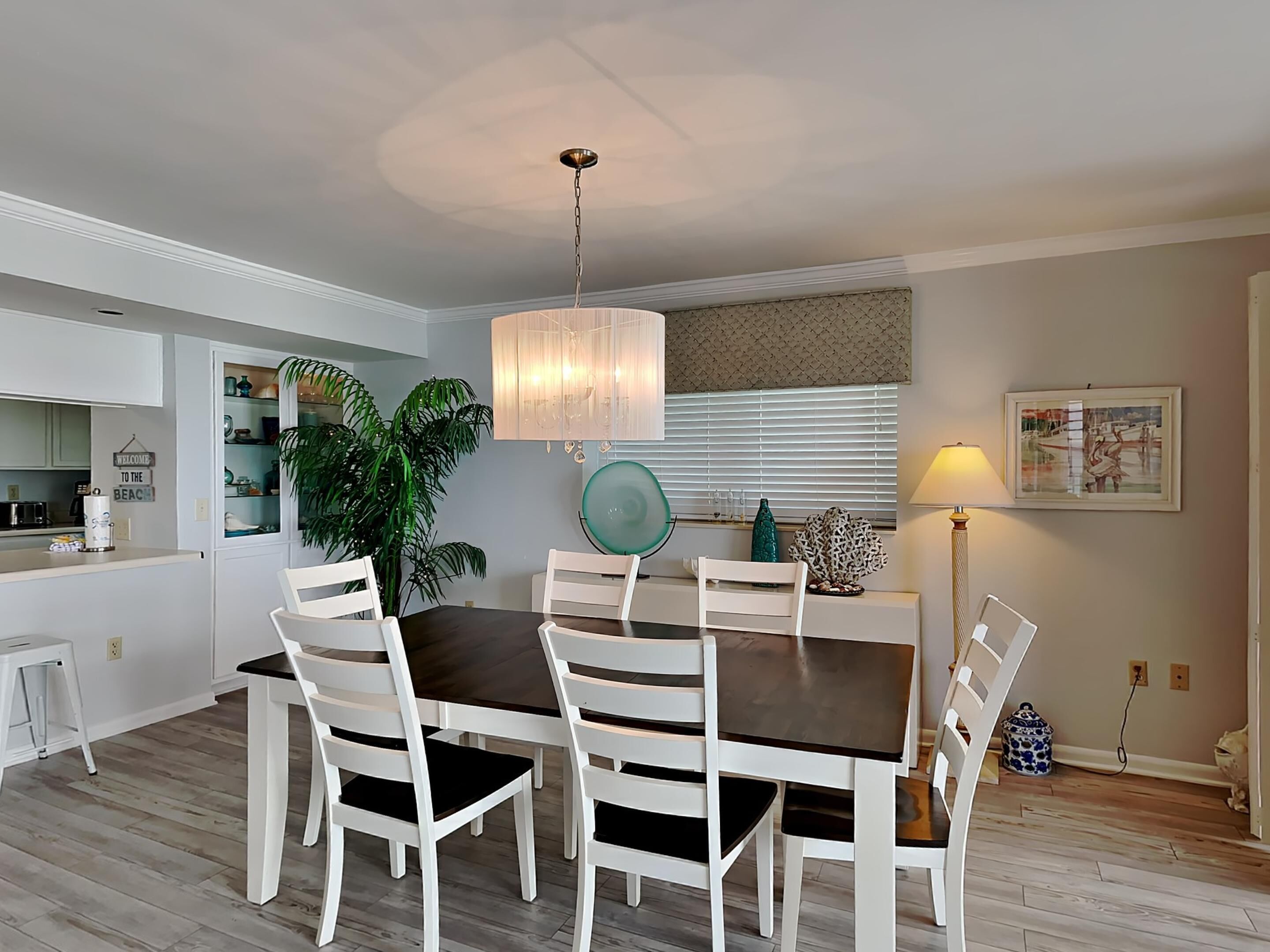 Un coin repas avec une table en bois foncé et des chaises blanches, un luminaire suspendu et une cuisine visible au fond.