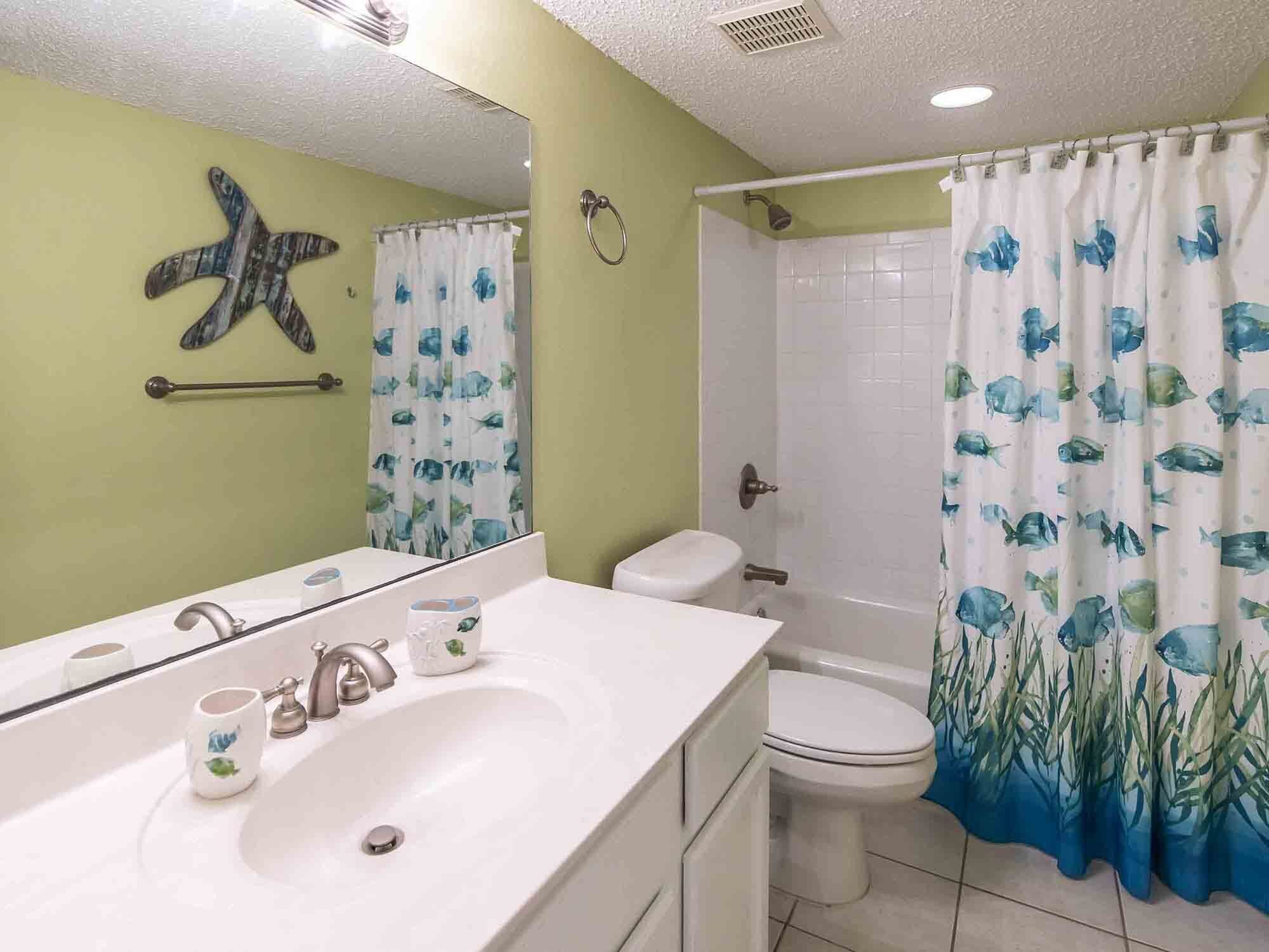 A bathroom with a white sink, a toilet, and a shower. There is a starfish decoration on the wall and a curtain with fish and seaweed patterns.