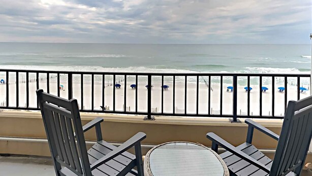 A balcony with two chairs and a small table, overlooking a beach with umbrellas and a railing. - Surf Dweller by Southern Vacation Rentals (Fort Walton Beach)