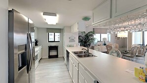 A modern kitchen with a stainless steel refrigerator, a long countertop, and a dining area with wicker chairs. - Surf Dweller by Southern Vacation Rentals (Fort Walton Beach)