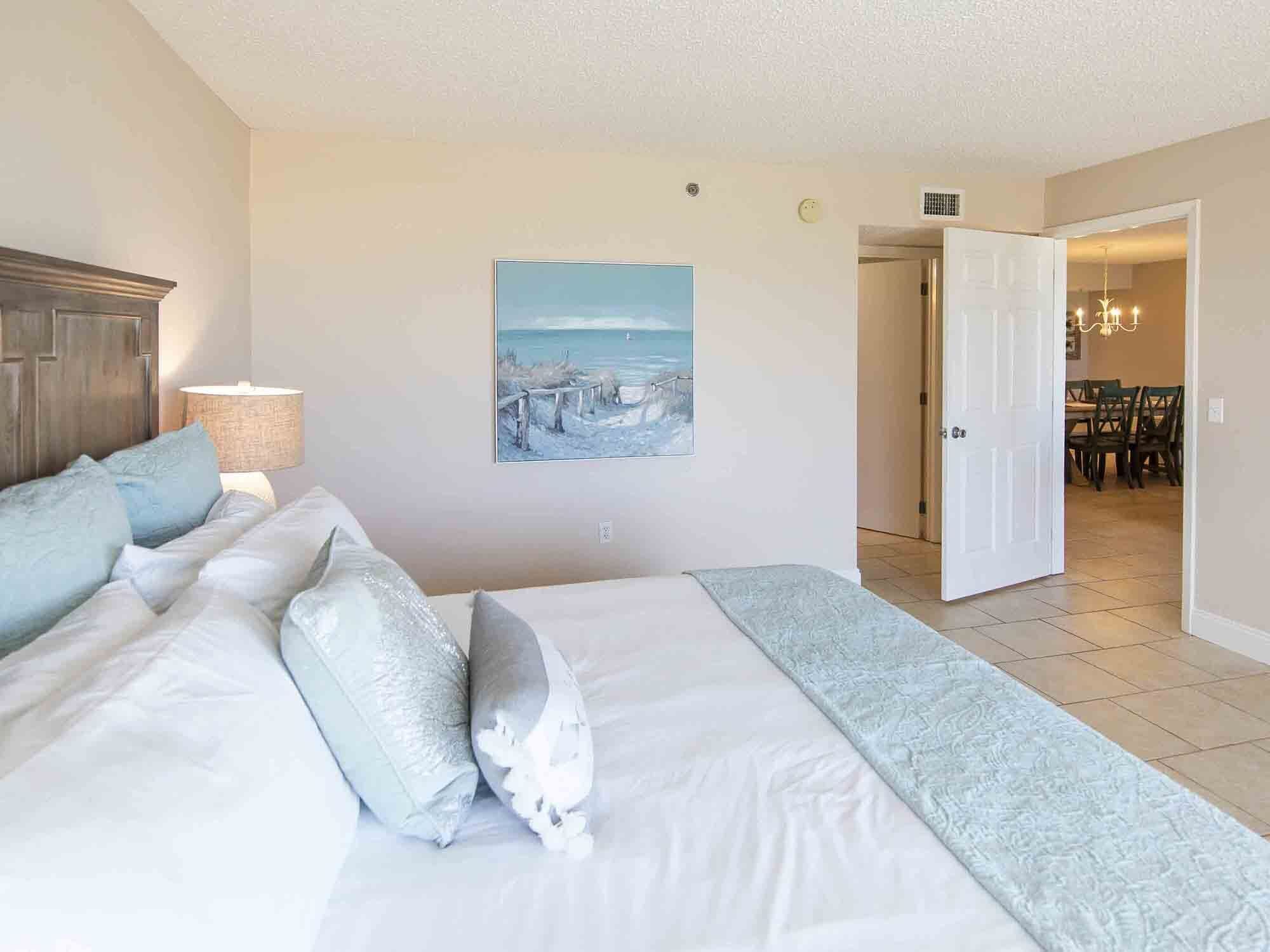 A bedroom with a large bed, a painting of a beach scene, and a view into a dining area.
