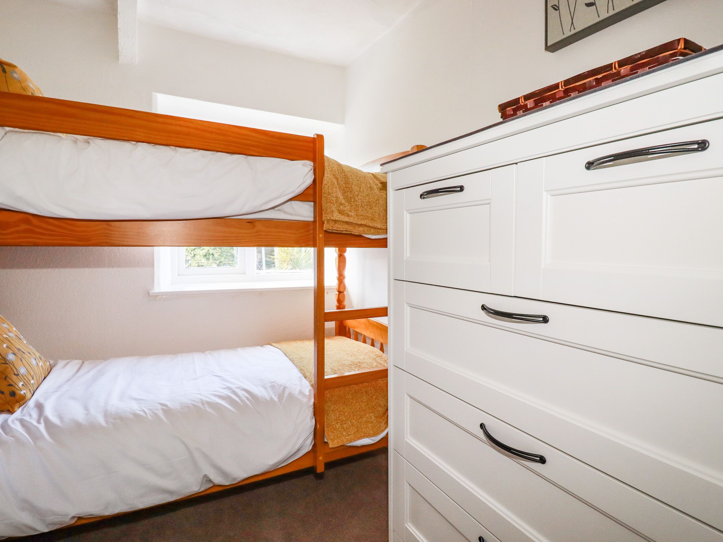 Una cama litera con ropa de cama blanca y un cabecero de madera, un aparador blanco con varios cajones, y una ventana que deja entrar luz natural.