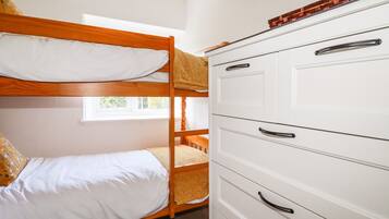 Una cama litera con ropa de cama blanca y un cabecero de madera, un aparador blanco con varios cajones, y una ventana que deja entrar luz natural.