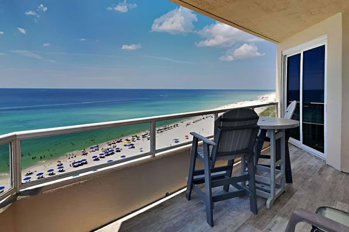 A balcony with a view of a beach, a table, and two chairs.