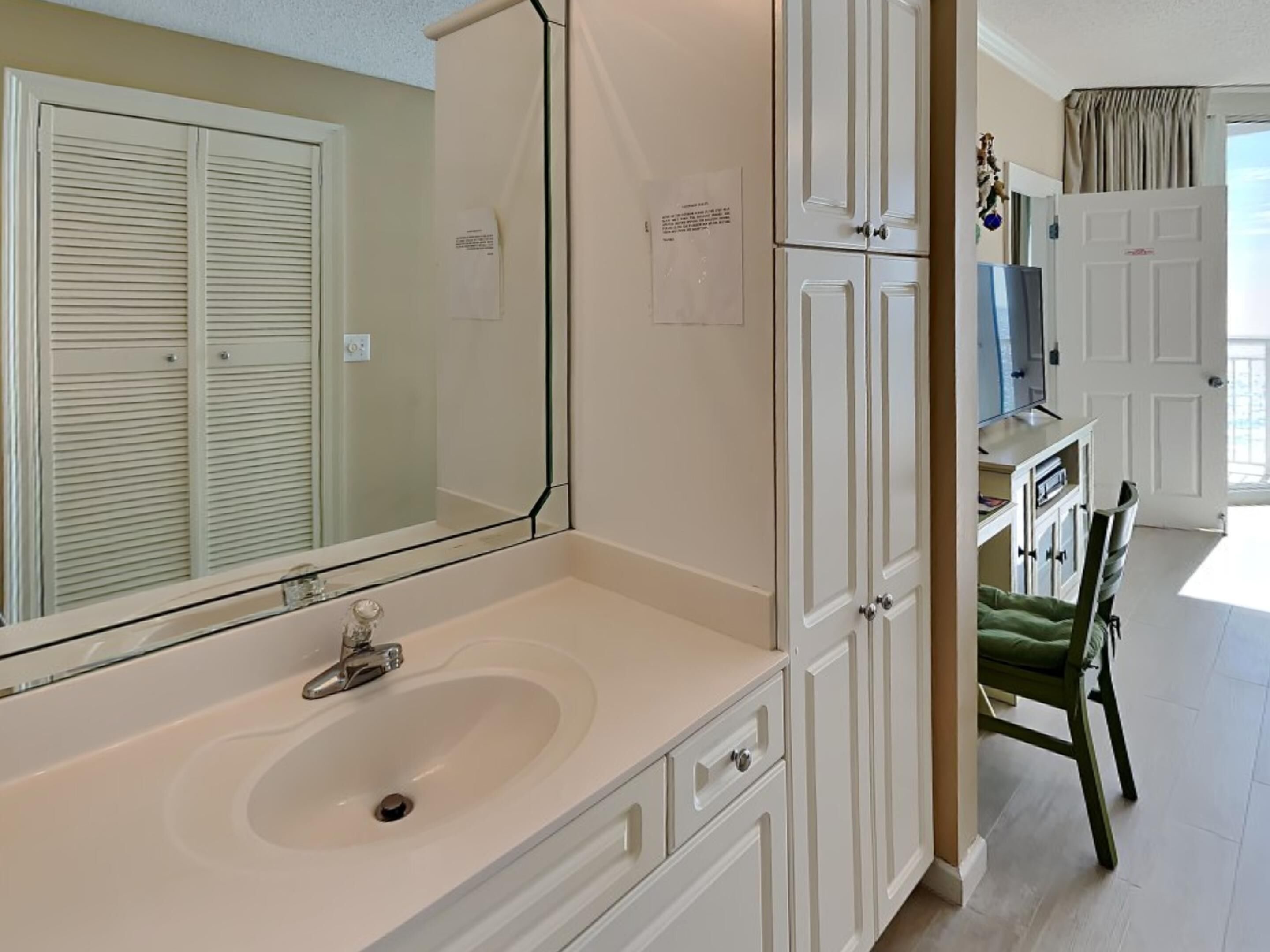A bathroom with a large mirror, white cabinets, a sink, and a chair.