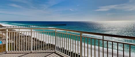 A wooden deck with a railing overlooking a beach and ocean, with a pier in the distance.