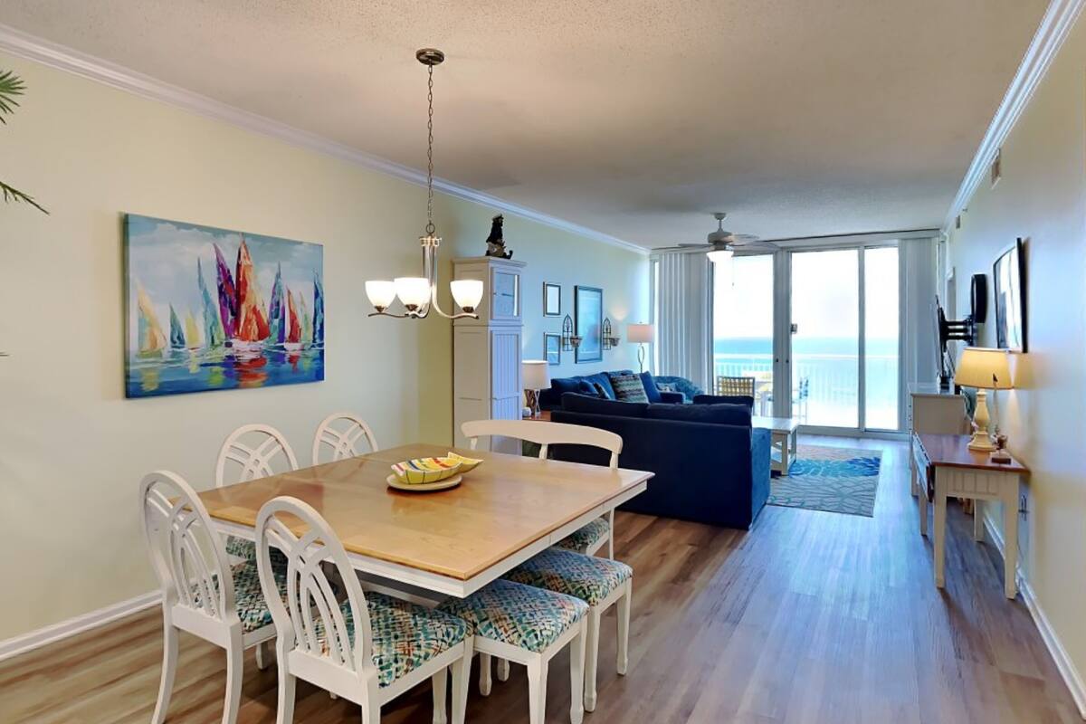 A dining area with a wooden table and white chairs, a living room with a blue sofa, and a view of the ocean through large windows.