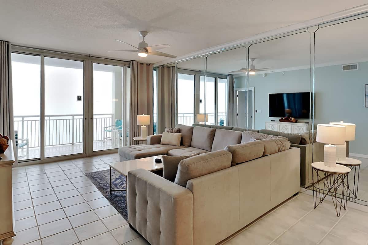 A modern living room with a large beige sectional sofa, a flat-screen TV, and a balcony with a view.