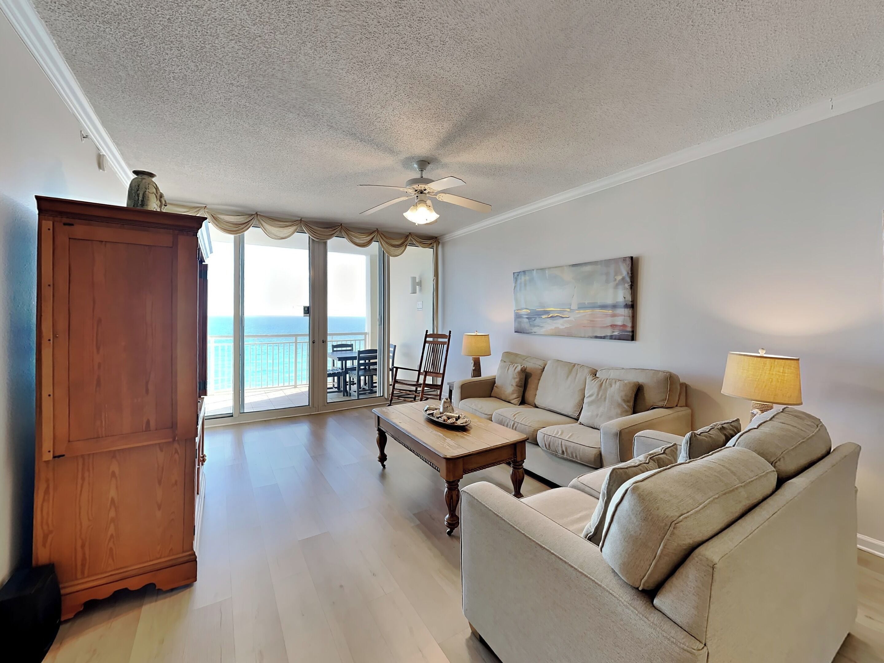 A spacious living room with a large sofa, a coffee table, and a dining table. There is a wooden cabinet and a ceiling fan. The room has a large sliding glass door leading to a balcony with a view of the ocean.