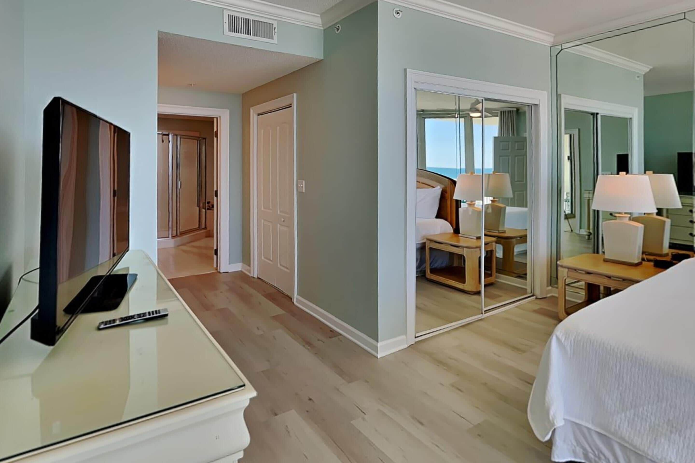 A hotel room with a bed, a desk with a laptop, a TV, and a mirror reflecting the room.